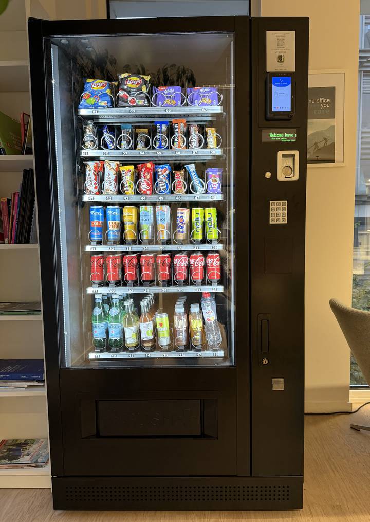Un distributeur installé dans un espace de bureau, sa vitrine éclairée montrant six rangées de snacks, de canettes et de bouteilles d’eau.
